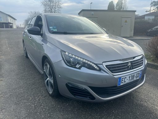 Peugeot 308 grise stationnée sur route, arrière d'un bâtiment en arrière-plan.